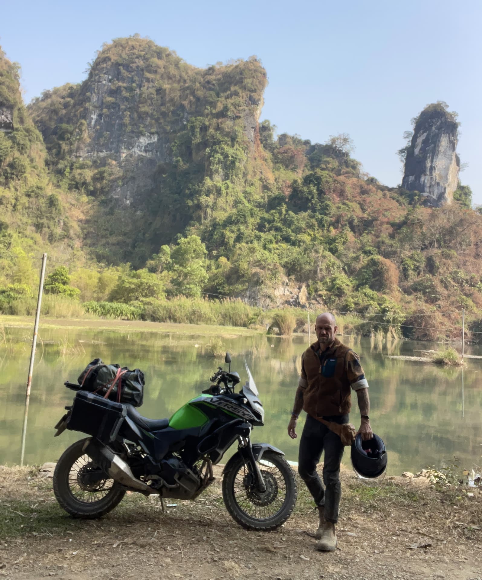 Jesse Kirley walking away from his loaded motorbike beside a river in Vietnam, limestone karst mountains behind